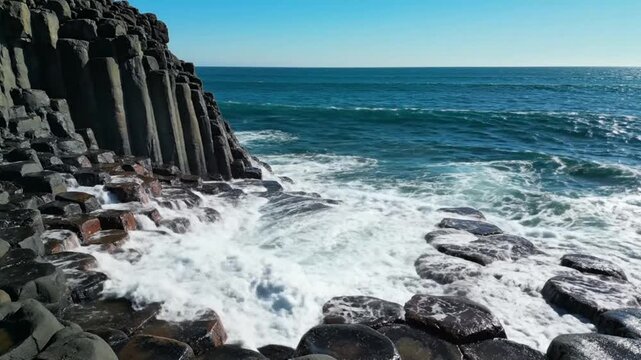 Waves crashing against hexagonal basalt rock formations on a sunny day at the coastline with blue ocean