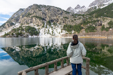 Woman standing on wooden viewpoint enjoying a calm mountain lake with perfect reflection in Panticosa, Pyrenees, Spain. Winter landscape with snow and peaceful atmosphere.