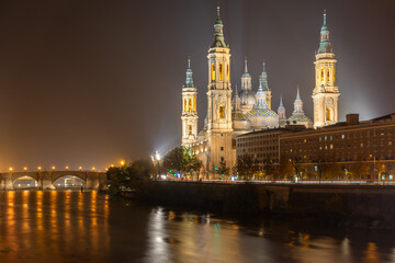 Fototapeta premium Night view of Basilica del Pilar illuminated with reflections on the Ebro river in Zaragoza, Spain. Iconic landmark with bridge and calm water in urban cityscape.