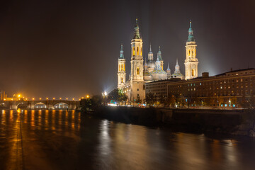 Fototapeta premium Night cityscape of Basilica del Pilar with light trails and reflections on the Ebro river in Zaragoza, Spain. Illuminated historic architecture and urban skyline.