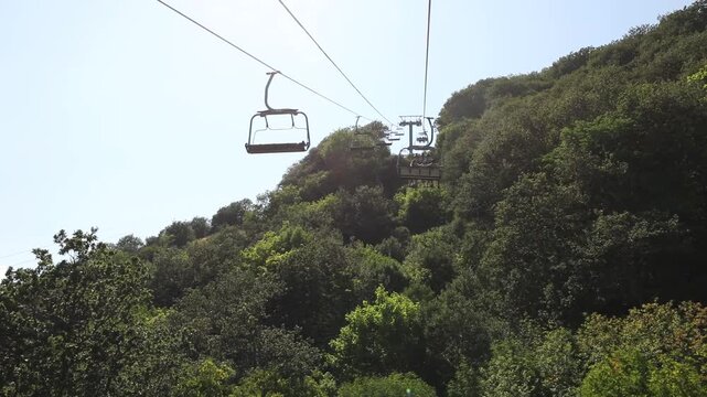 A scenic ski lift operates during summertime, transporting tourists up a mountain slope. The chairlift moves steadily over a dense lush green forest on a beautiful sunny day.