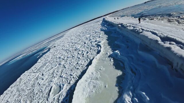 Aerial daytime view of frozen Baltic Sea at Daugava river mouth in Riga. Snow breakwater leads to lighthouse as people walk and ice drifts beside open water.