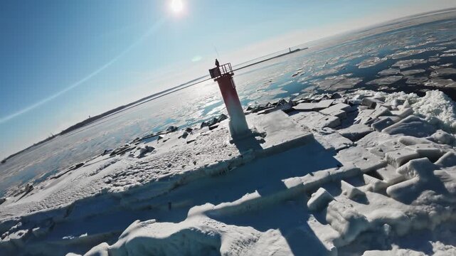 Aerial drone scene shows red lighthouse on Riga breakwater, Baltic Sea and Daugava mouth, blue water and white ice floes, tetrapod blocks with graffiti, midday light.