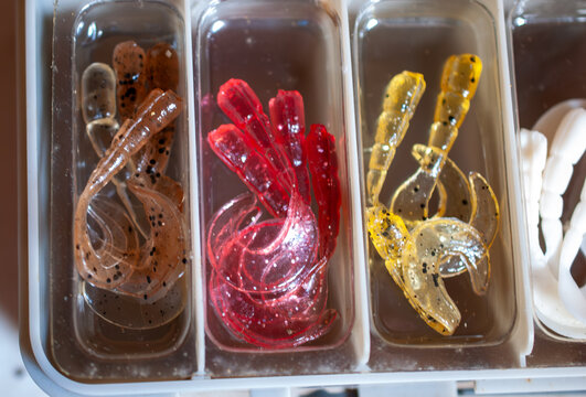 Assorted vibrant soft plastic fishing lures neatly organized in a tackle box, featuring red, brown, yellow, and white curly-tail grub baits with glitter for effective freshwater and saltwater fishing.