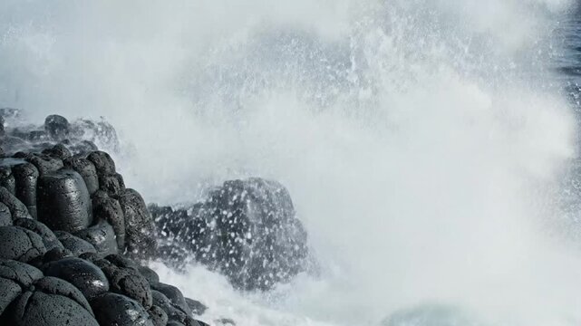 Powerful ocean waves crashing against black basalt rock formations, creating a dramatic coastal scene with splashing water