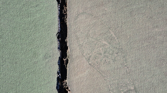 A top-down view of a green synthetic sports court divided by a wide, jagged vertical crack. The weathered, granular surface shows a faint, circular footprint mark on the right side of the fissure.
