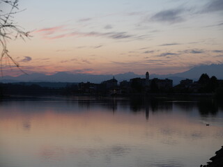Fototapeta premium Twilight view of Sesto Calende on Lake Maggiore, Italy. Warm lights reflecting on calm water, soft clouds and mountains in the background, serene atmosphere at dusk.