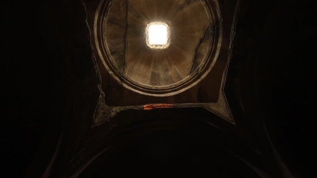 A camera slowly tilts down, revealing a beam of bright sunlight piercing through the central oculus of an ancient, dark, stone-carved dome inside a historic cave church interior.
