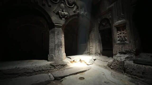 Walking through a dark doorway into the ancient rock-cut Geghard Monastery in Armenia. A shaft of light illuminates intricate stone carvings before revealing a dim room with candles.

