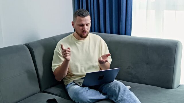 A man is sitting on a couch, using a laptop. He appears focused as he interacts with the computer. Sunlight enters the room through a window.