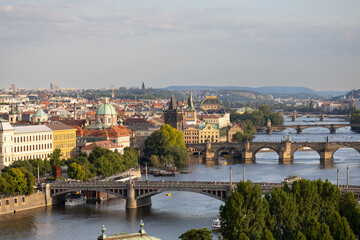 Obraz premium Aerial view of Prague cityscape featuring historic bridges, Vltava River, and colorful buildings under a clear sky with distant hills in the background
