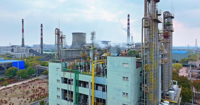 Industrial chemical plant and power station showing distillation columns, cooling towers, and smoke stacks with steam under a cloudy sky near an urban area.