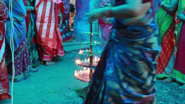 Close-up of a traditional multi-tier brass oil lamp (Deepam or Diya) illuminated during a Hindu religious ceremony in India. Surrounded by devotees in colorful sarees