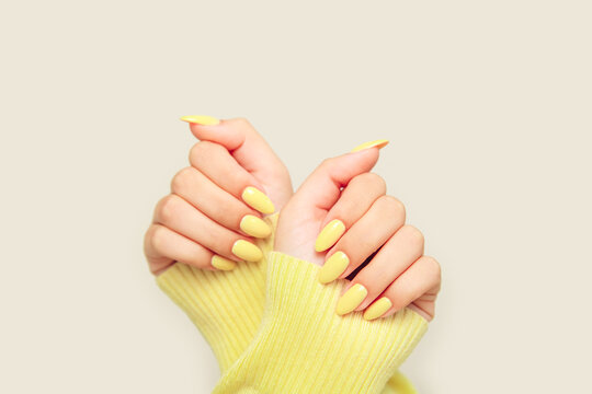 Close-up of female hands with pastel yellow nails wearing a yellow knitted sweater. Monochrome fashion aesthetic.