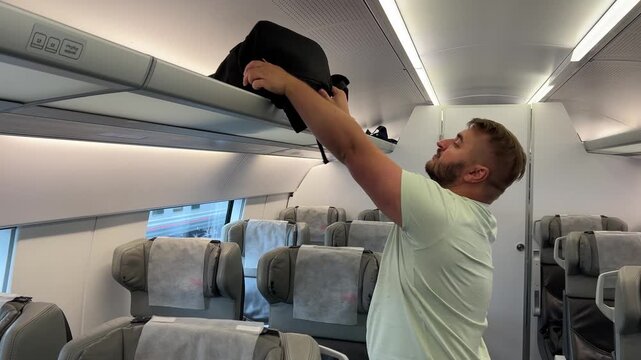 Horizontal 4K travel video. Young adult white man with beard and green t-shirt puts black backpack on baggage rack and sits on gray train seat. Summer vacation, tourism, arrival, transport, road trip