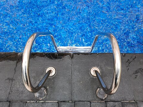 Close-up view of a swimming pool ladder with shiny metal rails and textured dark grey coping bordering the vibrant blue water