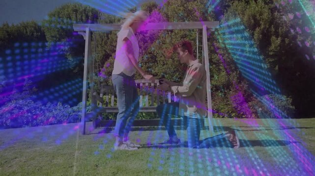 Vertical video: Kneeling man proposing placing ring on her hand after shared look by pergola, neon