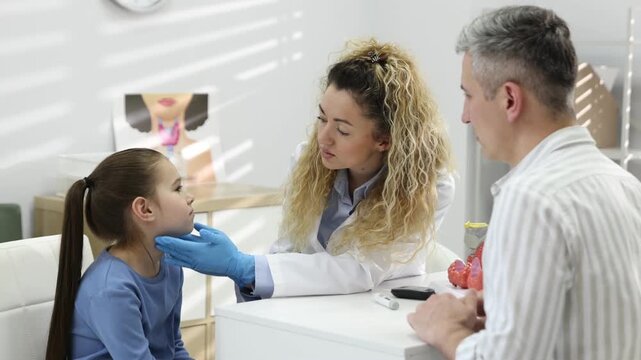 Endocrinologist in medical gloves examining girl's thyroid gland and her father at white desk in clinic. Camera moving forward