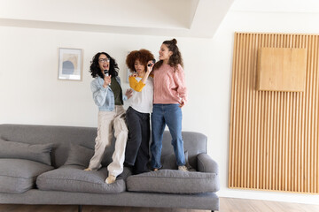 Diverse female friends standing on gray sectional sofa holding mics and singing in living room