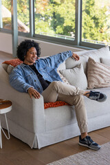 African American adult male sitting on light-gray sofa near large windows, smiling and leaning back