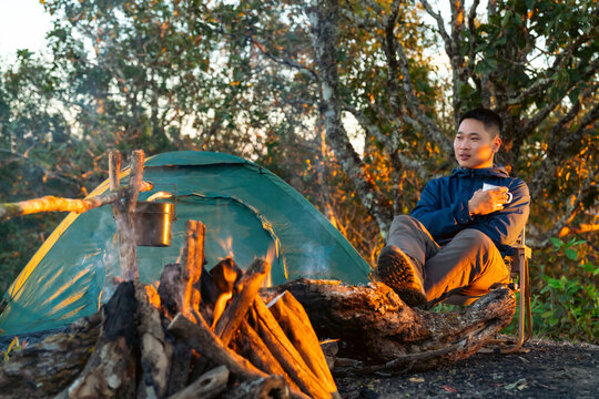 Asian man camping in forest drinking hot coffee beside tent and campfire, outdoor adventure active lifestyle, relaxing in nature during sunrise, social detox and solo travel wilderness experience