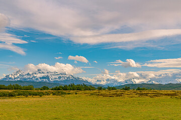 Obraz premium The iconic snow-covered peaks of the Paine Massif rise above green grasslands under blue sky with scattered clouds at Torres Del Paine National Park, Chilean Patagonia.