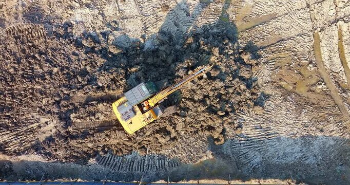 Top down aerial view of yellow excavator working on muddy construction site, moving earth for infrastructure development and land preparation.