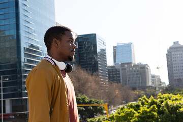 Fototapeta premium African American man in 20s standing left on plaza wearing brown jacket and headphones, copy space