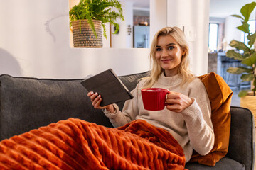 Adult woman reclining in living-room sofa snuggling rust blanket holding tablet folio and red mug