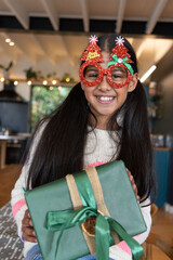 Asian child girl smiling and holding green-wrapped gift in kitchen wearing glittery red glasses