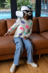 African American girl child sitting on brown leather sofa wearing VR headset reaching toward pool