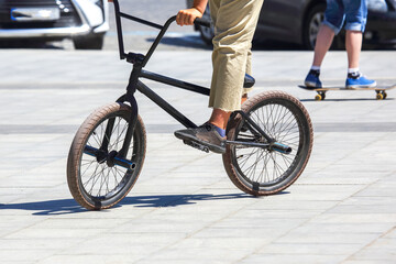 Naklejka premium Biker rides a black BMX bike on pavement while a person skateboards in the background during a sunny day
