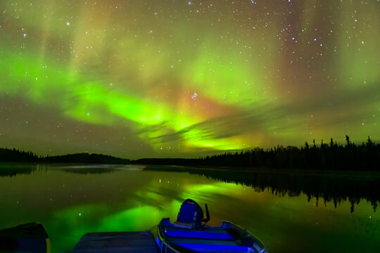 Green aurora borealis dances over calm lake with docked boats and wooden pier in foreground
