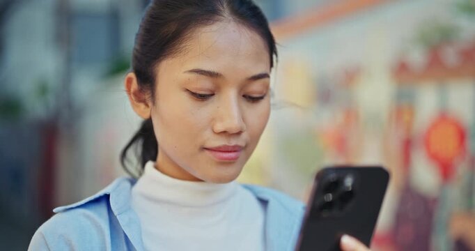Smooth slow motion video of a cheerful young Asian woman smiling at her smartphone screen while interacting with an AI assistant outdoors with natural light.