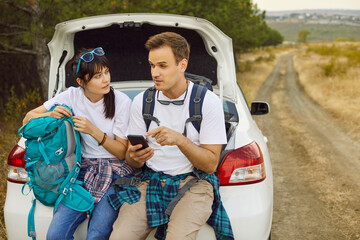 Fototapeta premium Couple by car use phone, map, backpack. Stranded on a rural road, travelers check geolocation, seek signal, and study a map while resting on trunk near forest. Adventure on weekend travel and hiking.