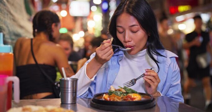 Young Asian woman enjoying a traditional meal at a roadside food stall in a busy night market with bokeh lights and people in background.