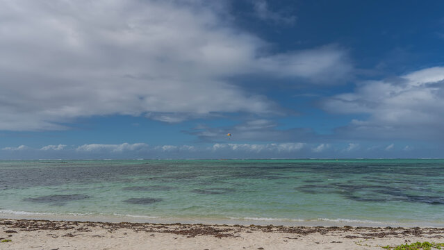 A calm turquoise ocean. A boat is visible in the distance. A multicolored parachute is floating in the blue sky among the clouds. Dry algae on a sandy beach. Mauritius. &Icirc;le aux Cerfs 