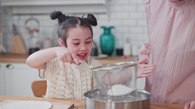 Happy and cheerful young Asian girl smiles while using a sieve to sift flour into a pot with her mother's guidance during a fun home baking session.