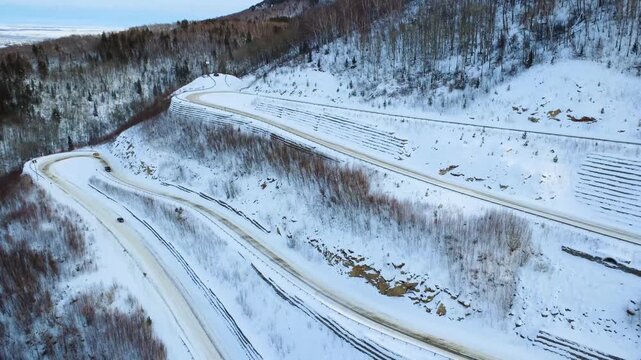 Aerial View Engineered Mountain Road Cut Into Hillside With Embankments And Cleared Lanes, Visible Plow Marks And Tracks In Snow, Infrastructure Contrast With Surrounding Forest, Ideal