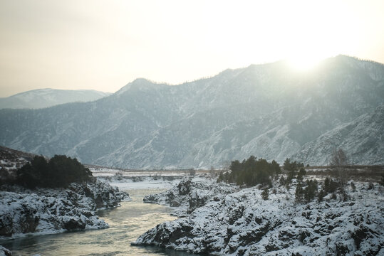 Sunlit Snowy River Winding Through Mountain Valley, Soft Golden Light, Gentle Mist Rising From Water, Snowdusted Pines Along Banks, Peaceful Atmosphere And Crisp Cold Air Capturing Tranquil Dawn.