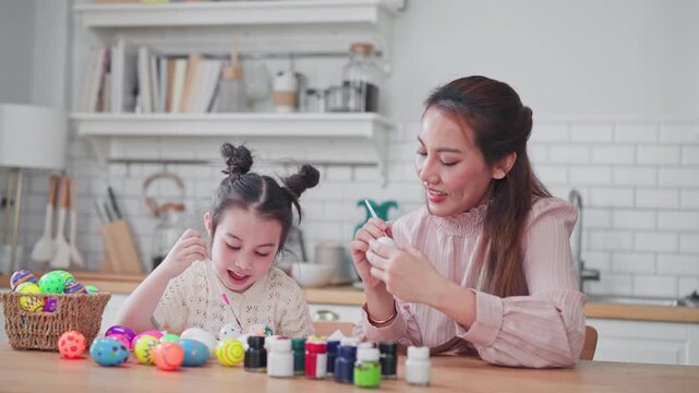 Loving mother ties her daughter's hair during a painting session. The mother and child enjoy painting colorful Easter eggs together for a fun family holiday in their kitchen.
