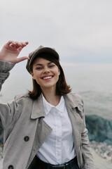 Smiling young woman in casual coat and hat stands near rocky seaside, enjoying fresh air and...