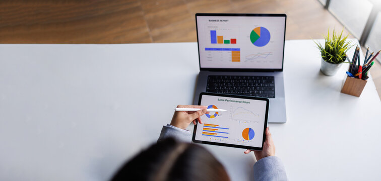 A bird's-eye view of a professional examining sales performance data on a tablet, while also considering a laptop report, highlighting the importance of analytics in business.