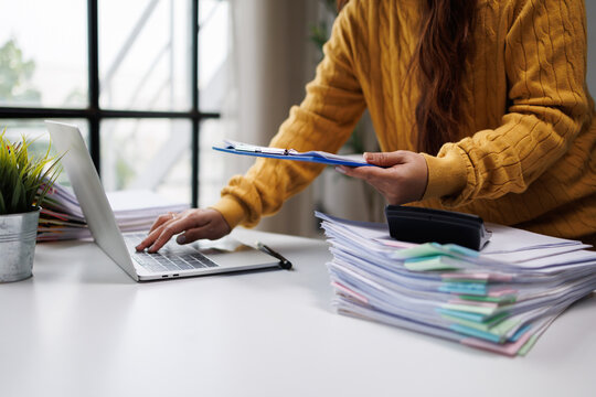 A woman balances her laptop usage with organizing stacks of colorful paperwork, blending modern technology with traditional office tasks in a bright environment.