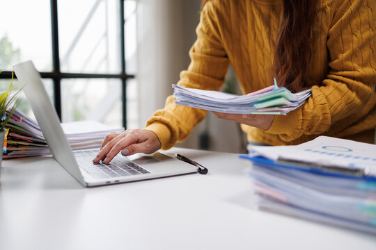 A woman in a yellow sweater organizes several stacks of documents while using her laptop, representing an engaging work atmosphere in a modern office setup.