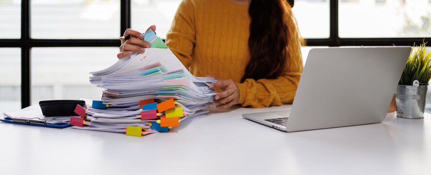 A woman efficiently organizing a tall stack of documents with colorful tabs while working on her laptop in a bright, modern office space filled with natural light.