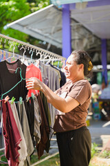 Hanging laundry to dry outdoors instead of using a dryer. Mature Asian woman hangs laundry outside sunny backyard. Housewife organizes wet clothes hanging metal rack.