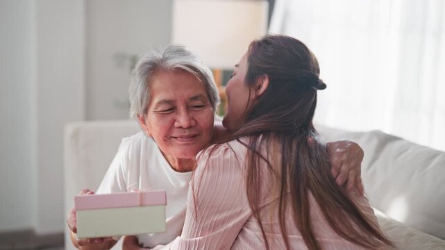 Happy Asian daughter presents a Mother's Day gift to her loving mother. The heartwarming mother and child celebrate with a handwritten card and a gift box in their home.