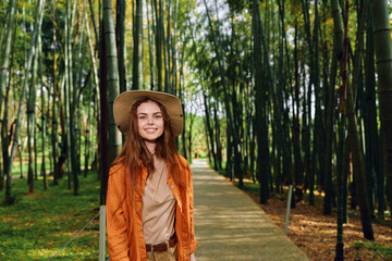 Fototapeta premium Woman smiling in straw hat and orange jacket walking on wooden path through bamboo forest, portrait of traveler in nature with sunlight filtering through tall bamboo stalks.