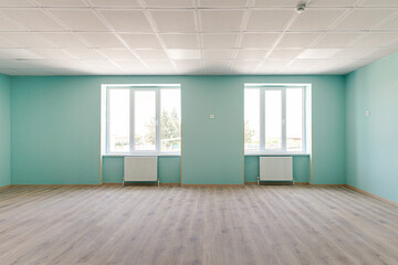 Fototapeta premium Interior view of a spacious empty hall featuring bright turquoise walls, two large windows with radiators, wooden laminate flooring and a professional acoustic grid ceiling.